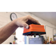 A hand holding an orange and black magnetic pop up tray upside down with three metal screws visible, set against a blurred workshop background to show the screws are not able to be tipped out
