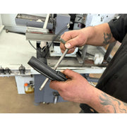 A close-up of a person's hands using a metal tool to work on a black cylindrical component in an industrial workshop setting.