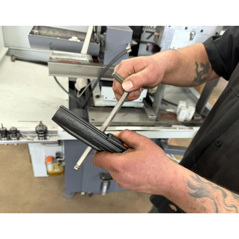 A close-up of a person's hands using a metal tool to work on a black cylindrical component in an industrial workshop setting.