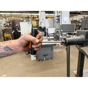 A close-up of a person's tattooed hand holding a black cylindrical tool with an orange accent, positioned near a metal clamp in a workshop or industrial setting.