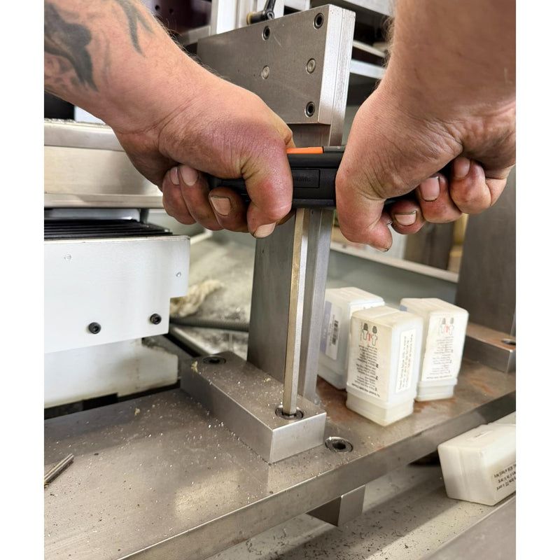 Close-up of a person's hands operating a metal clamp or press on a workbench, with small white rectangular product blocks visible in the background.