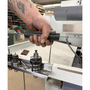 A close-up of a person's tattooed hand holding a black handheld tool against a white and gray industrial machine in a workshop setting.