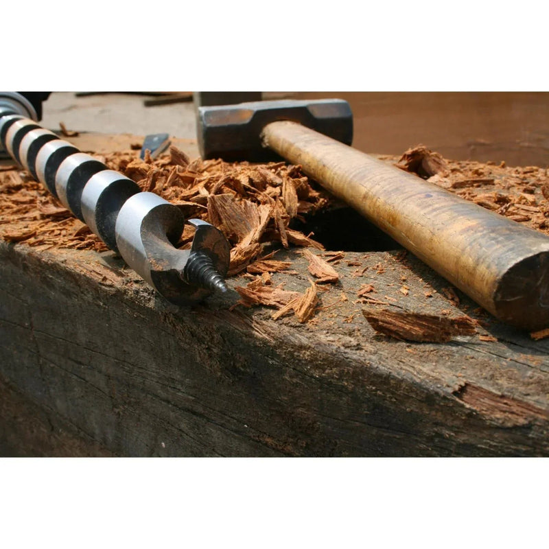 Wooden mallet and metal auger on a wooden surface with wood shavings.